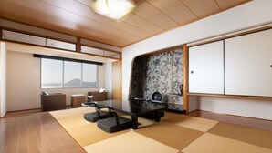 Una habitación amplia con suelo de tatami, una mesa baja con cojines, un ventanal con vistas y una puerta corredera con un panel decorativo.