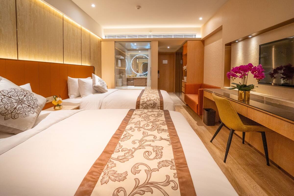 A hotel room with two beds, a desk, a chair, a mirror, and a vase with flowers.