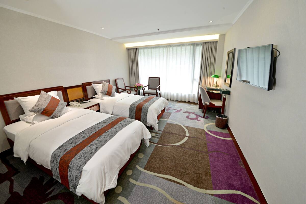 A hotel room with two beds, a desk, a chair, a television, and a large window with curtains.