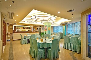 Une salle à manger d’hôtel avec des tables rondes, des chaises recouvertes d’un tissu vert et un luminaire central.
