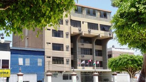 A multi-story building with a central entrance, surrounded by a white fence and a blue building to the left. - Intiotel - Chiclayo (Chiclayo)