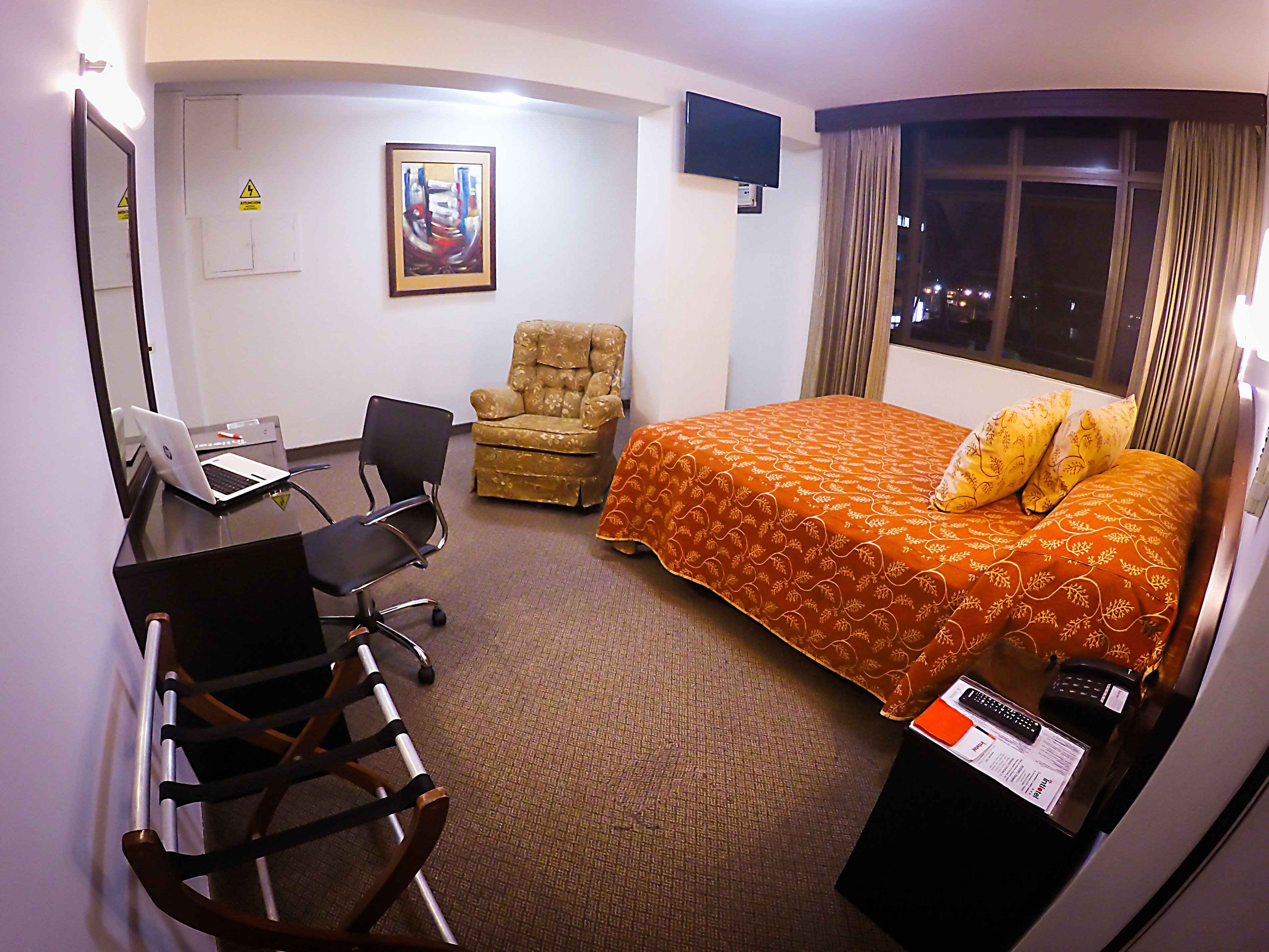 Une chambre d’hôtel avec un lit, un bureau avec un ordinateur portable, une chaise, une télévision et une fenêtre avec des rideaux.