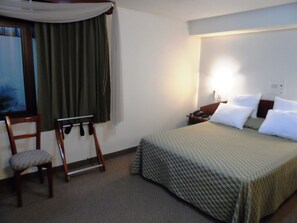 A hotel room with a bed, a chair, a nightstand, and a window with curtains. - Intiotel - Chiclayo (Chiclayo)