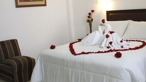 A neatly made bed with white linens, a striped armchair, and a vase with red roses on the side. - Intiotel - Chiclayo (Chiclayo)
