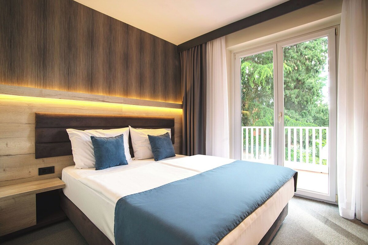A hotel room with a large bed, a wooden headboard, a nightstand, and a balcony with a white railing.