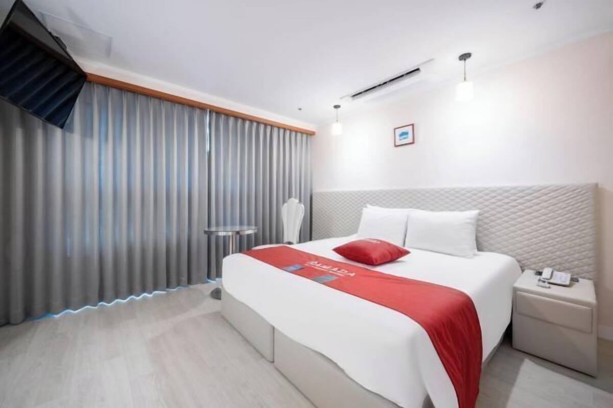 A hotel room with a bed, a nightstand, a wall-mounted air conditioner, and a window with blinds.