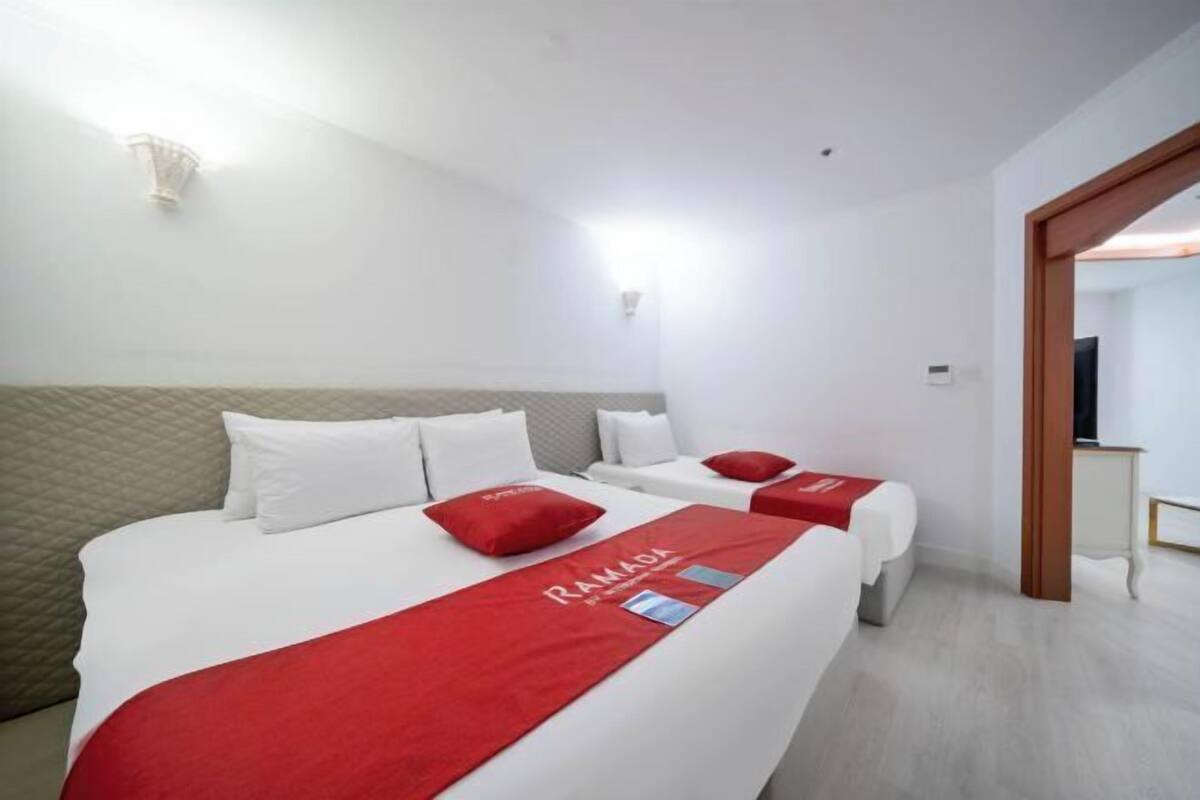 A hotel room with two beds, a red bed runner, a white headboard, and a wall-mounted light fixture.