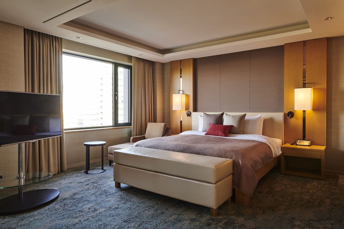 A modern hotel room with a large bed, a flat-screen TV, a small round table, and a bench.