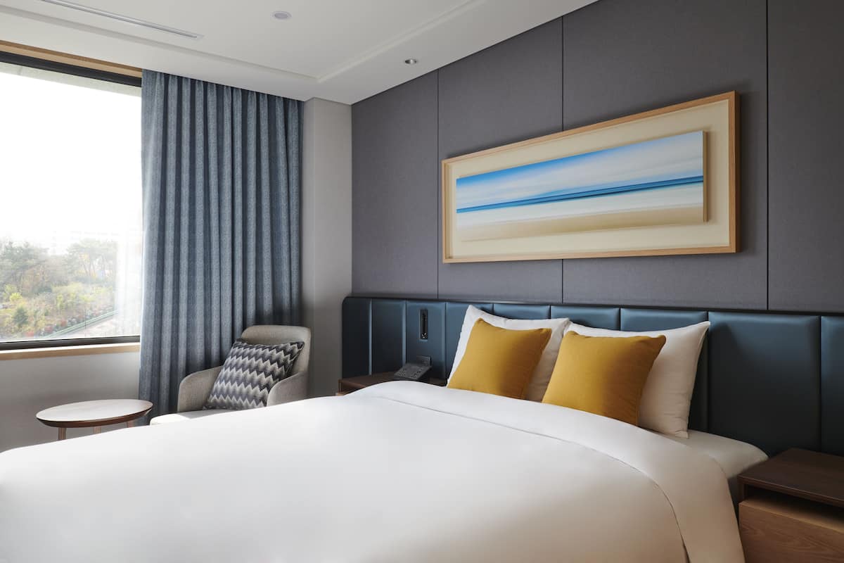 A modern hotel room with a large bed, a bedside table, a chair, and a painting on the wall.