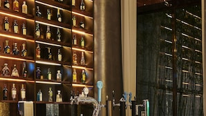 Una zona de bar con varias estanterías llenas de botellas, una barra y cuatro taburetes de bar.