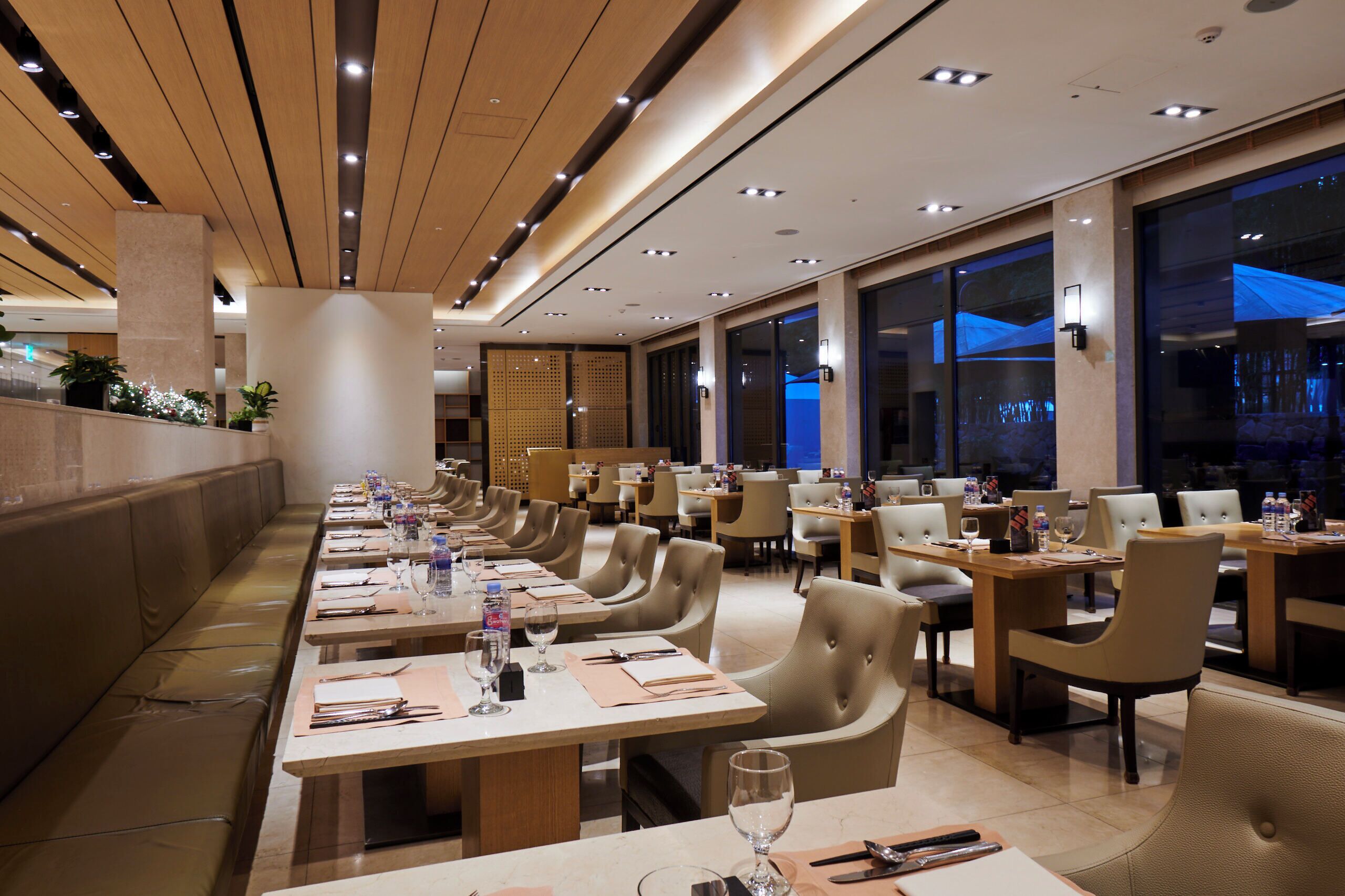 A modern dining area with multiple tables and chairs, a long bench, and a view of the night sky through large windows.