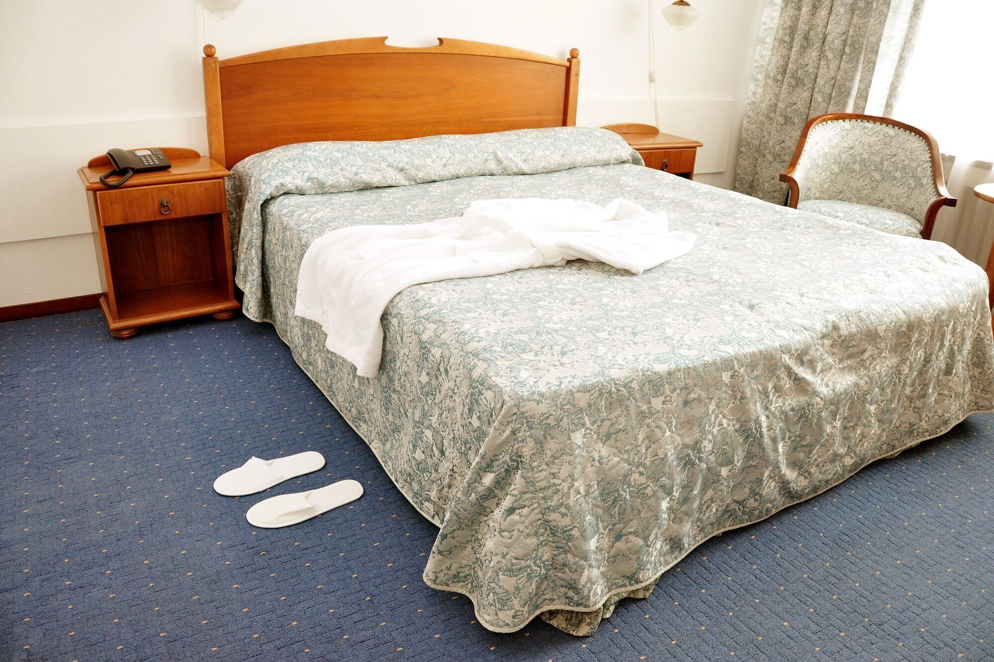 A neatly made bed with a floral bedspread, a wooden headboard, a bedside table with a telephone, and a chair with a floral pattern.