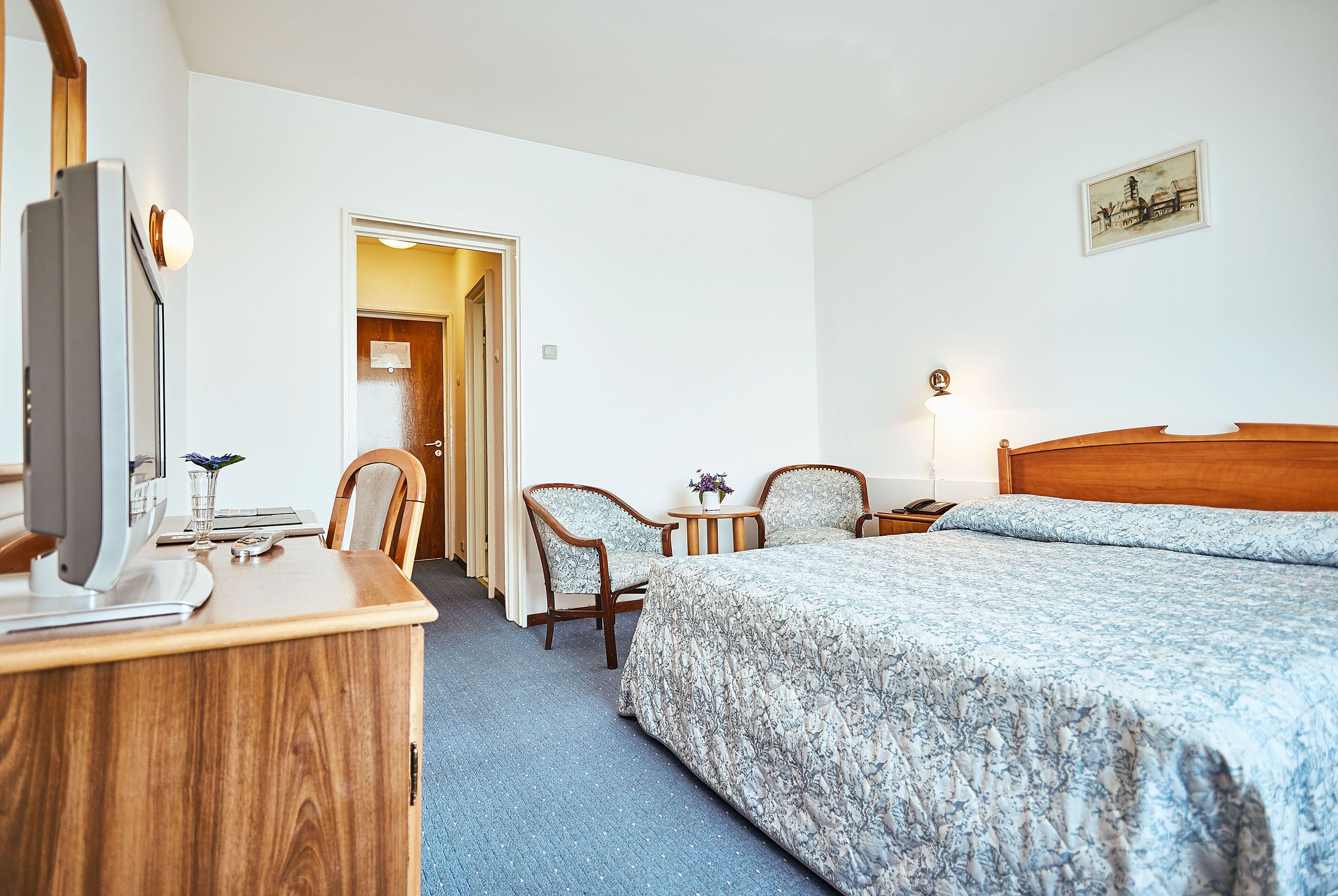 Ein Hotelzimmer mit Holztisch, zwei Stühlen, einem Bett mit floralem Bettzeug, einem Nachttisch und einer Wandlampe.