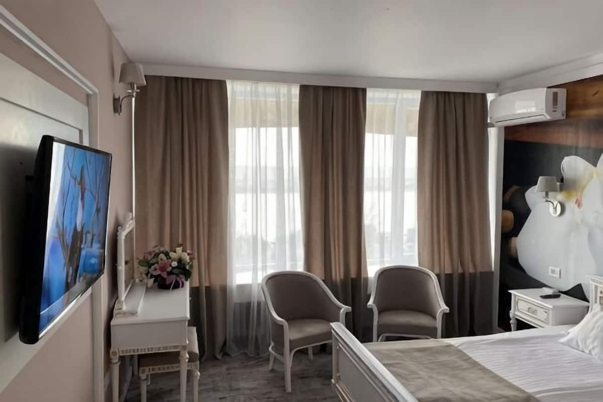 Quarto de hotel com cama, duas cadeiras, escrivaninha, televisão e uma janela grande com cortinas.