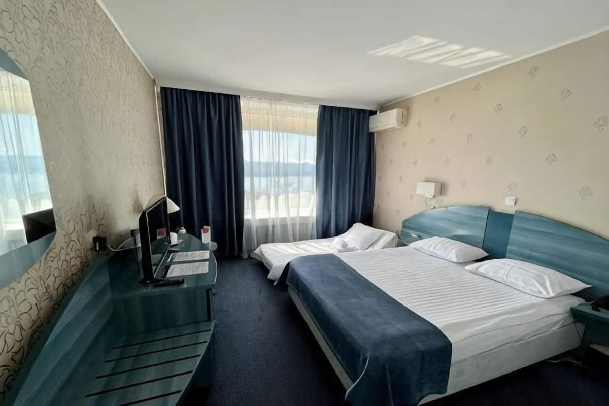Quarto de hotel com duas camas, uma mesa com computador, janela com cortinas e unidade de ar condicionado.