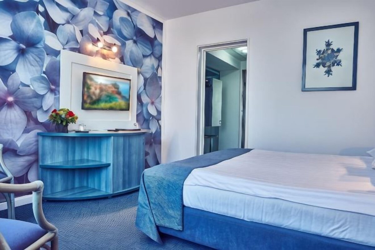 Une chambre d’hôtel avec un lit, un téléviseur posé sur une console bleue, un tableau encadré au mur et une porte donnant sur une autre pièce.