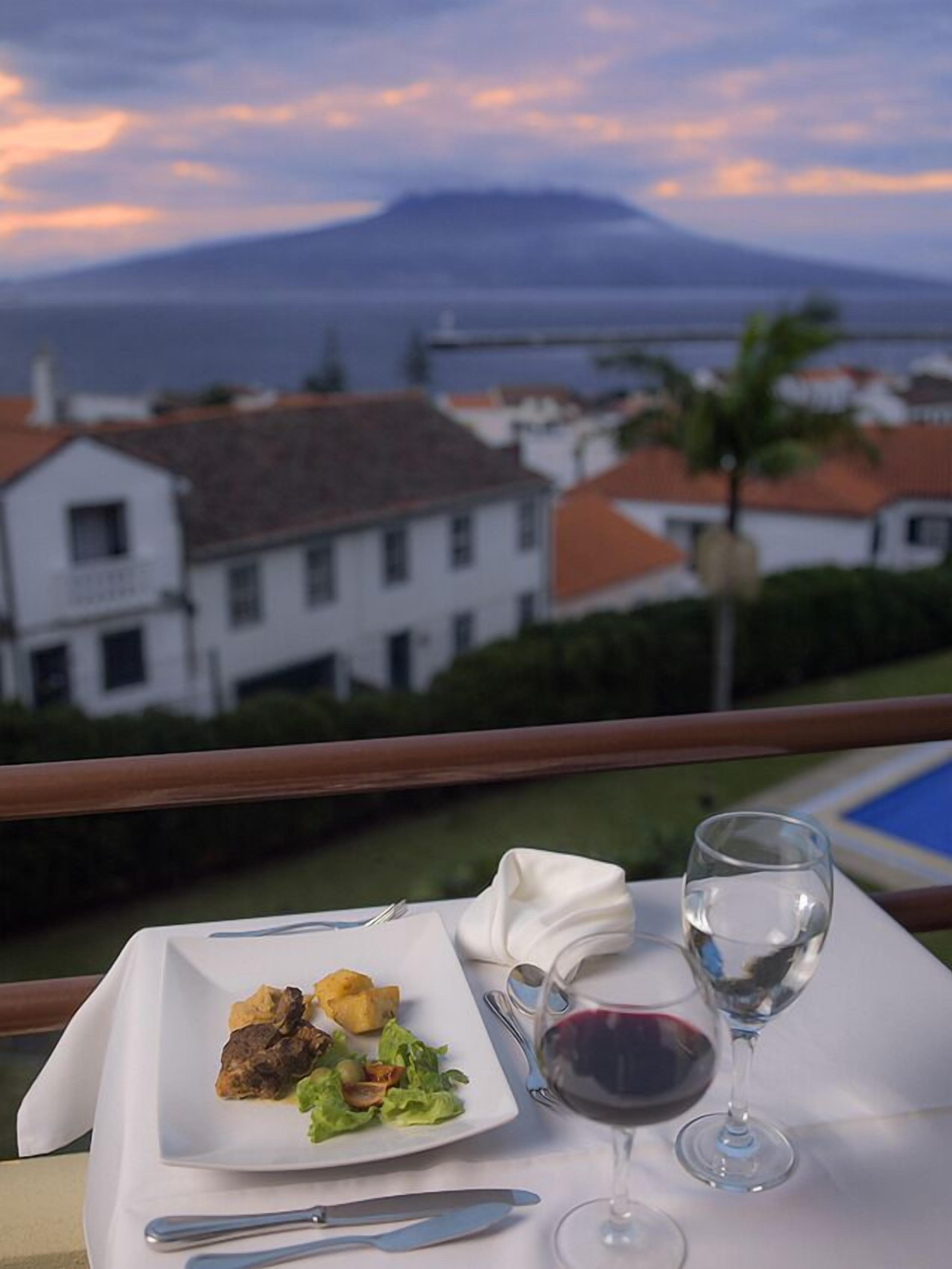 A dining table with a plate of food, a glass of red wine, and a view of a coastal town at sunset.