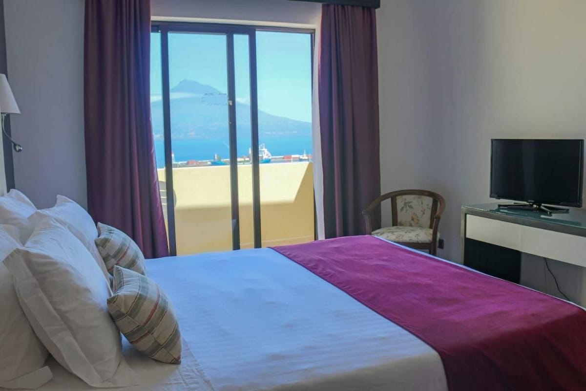 A hotel room with a bed, a television, a chair, and a view of the sea and mountains through a sliding door.