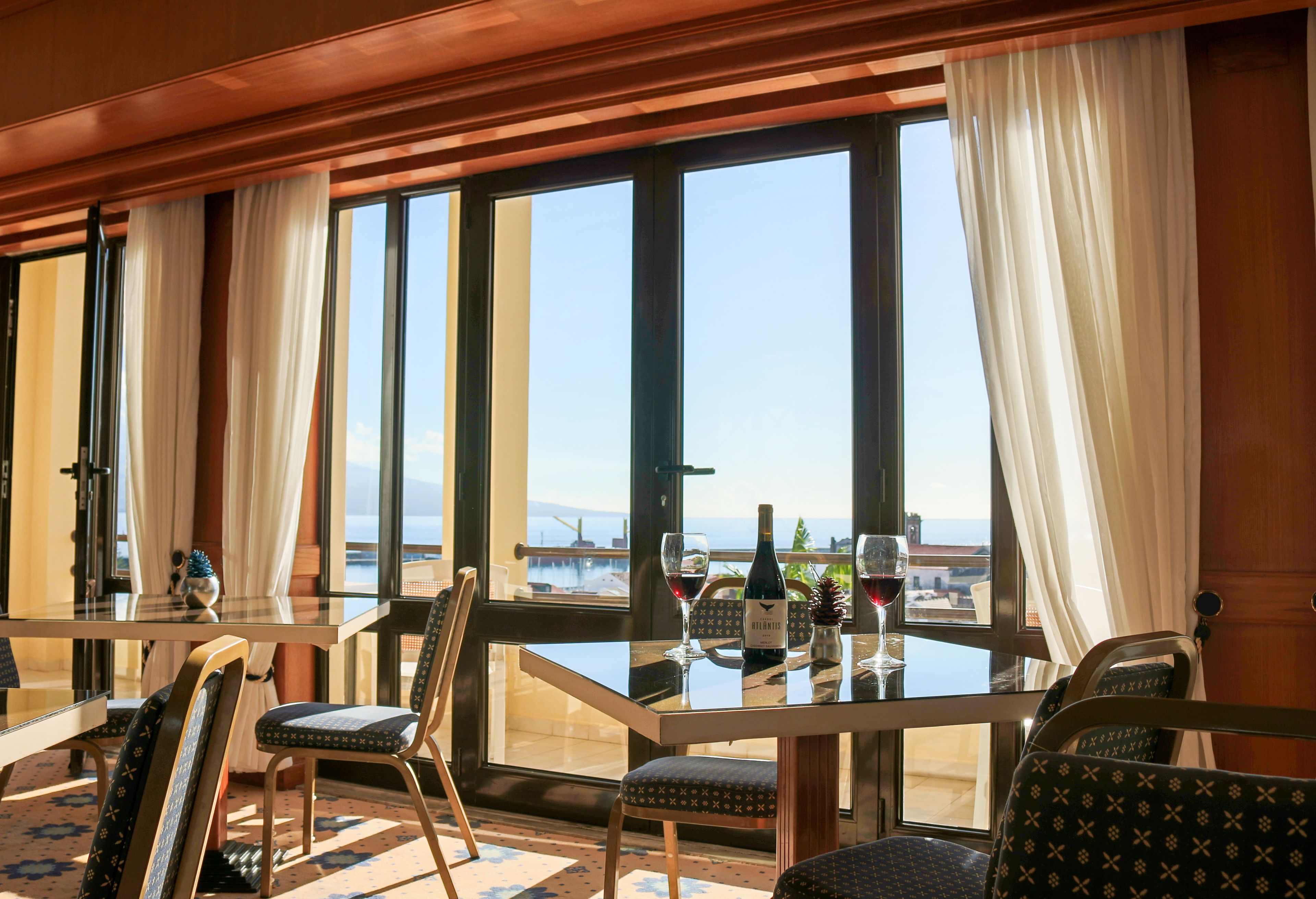 Ein Essbereich mit einem Tisch für zwei Personen, großen Fenstern mit Vorhängen und Blick auf das Meer.