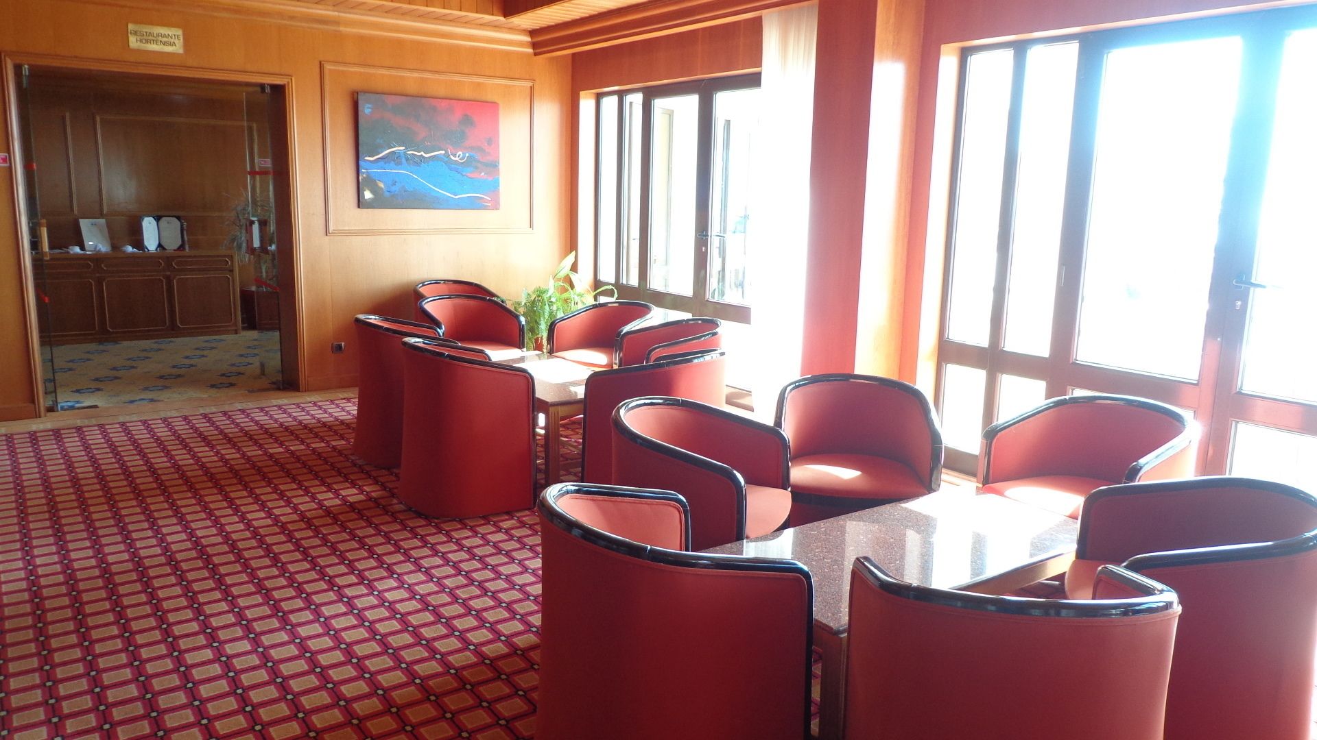 A dining area with a table and red chairs, a painting on the wall, and large windows.