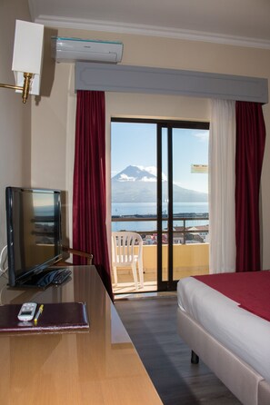 A hotel room with a bed, a desk with a TV and phone, a view of a mountain and sea through a balcony. - Hotel Horta (Horta)