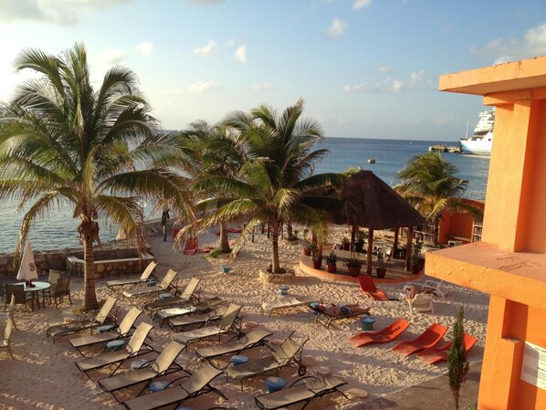 A beachside hotel with sun loungers, palm trees, and a thatched-roof structure.