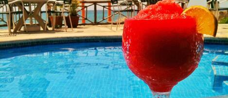 Escena junto a la piscina con un vaso de bebida roja, sillas de piscina y sombrillas.