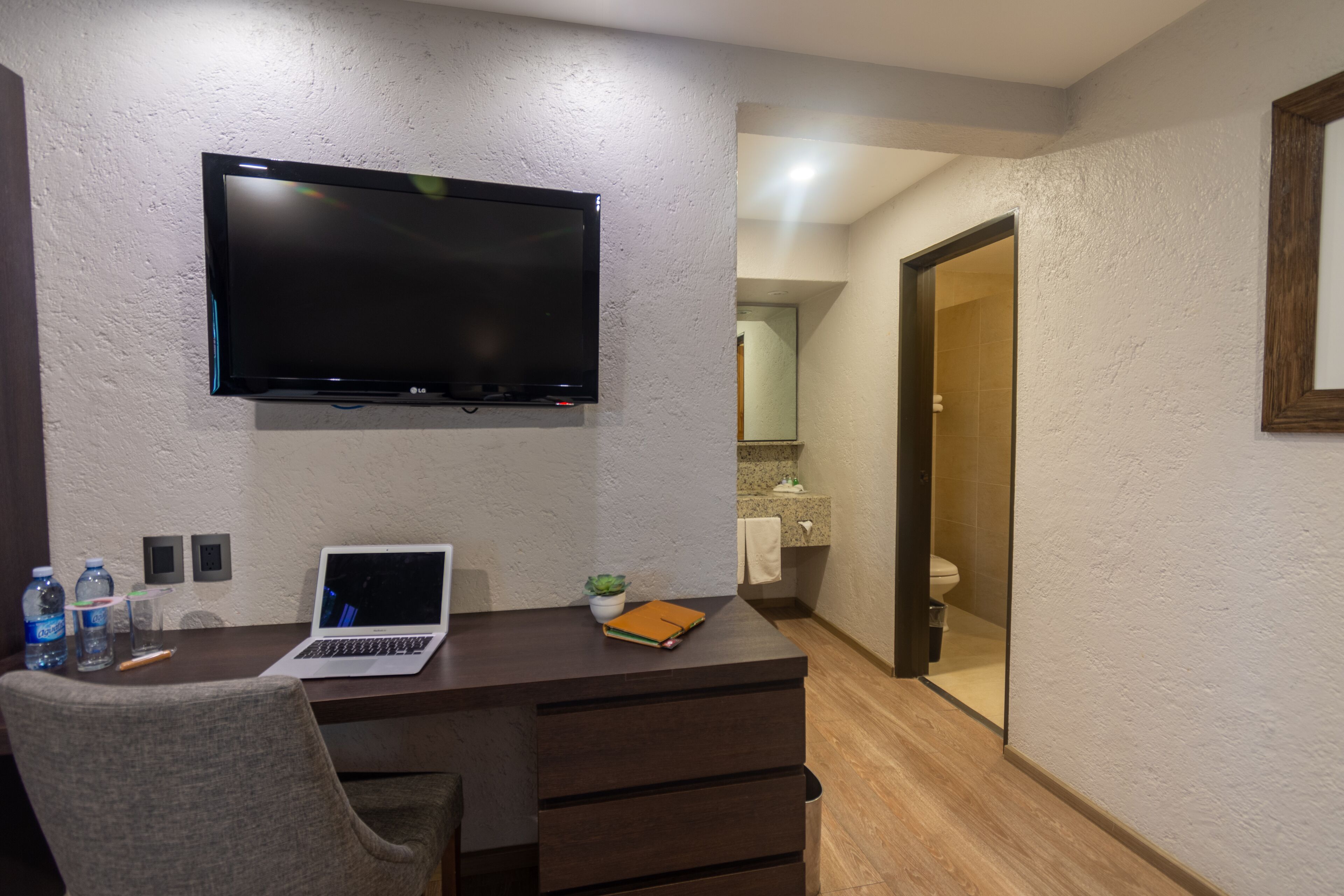 Hotelkamer met een bureau, laptop, flatscreen televisie, stoel, een kleine plant en een badkamer zichtbaar door een geopende deur.