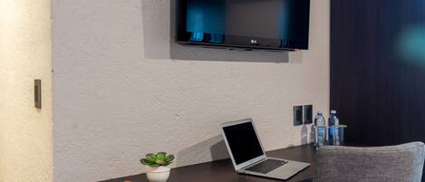 Una habitación moderna con televisión de pantalla plana en la pared, una laptop en un escritorio, una pequeña planta en maceta, una libreta amarilla y una silla gris.