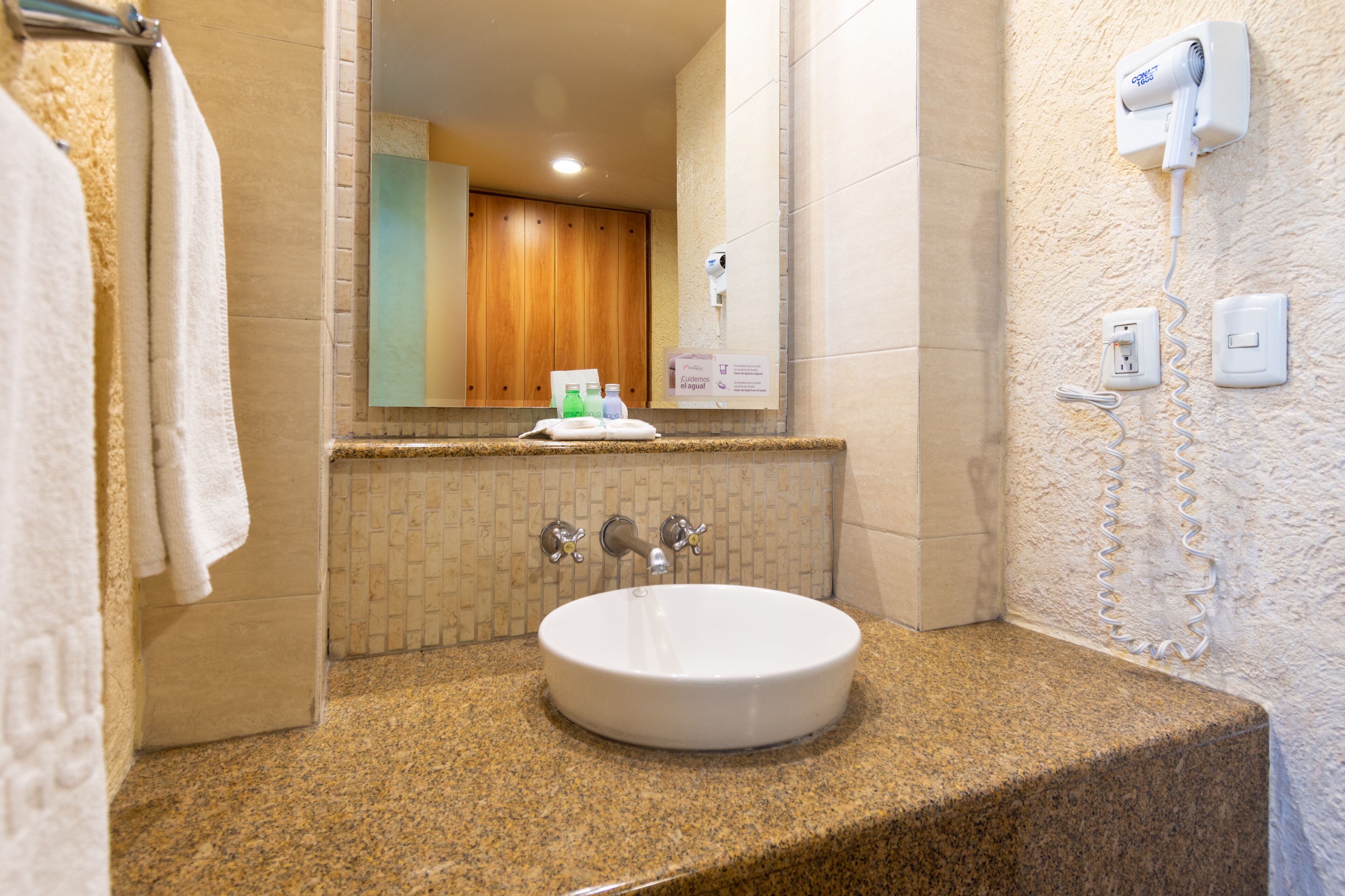 Un baño con encimera de granito, un lavabo blanco, un espejo, un perchero, un secador de pelo y un enchufe en la pared.