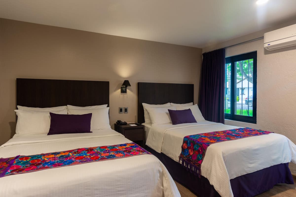 A hotel room with two beds, a nightstand with a lamp, and a window with curtains.