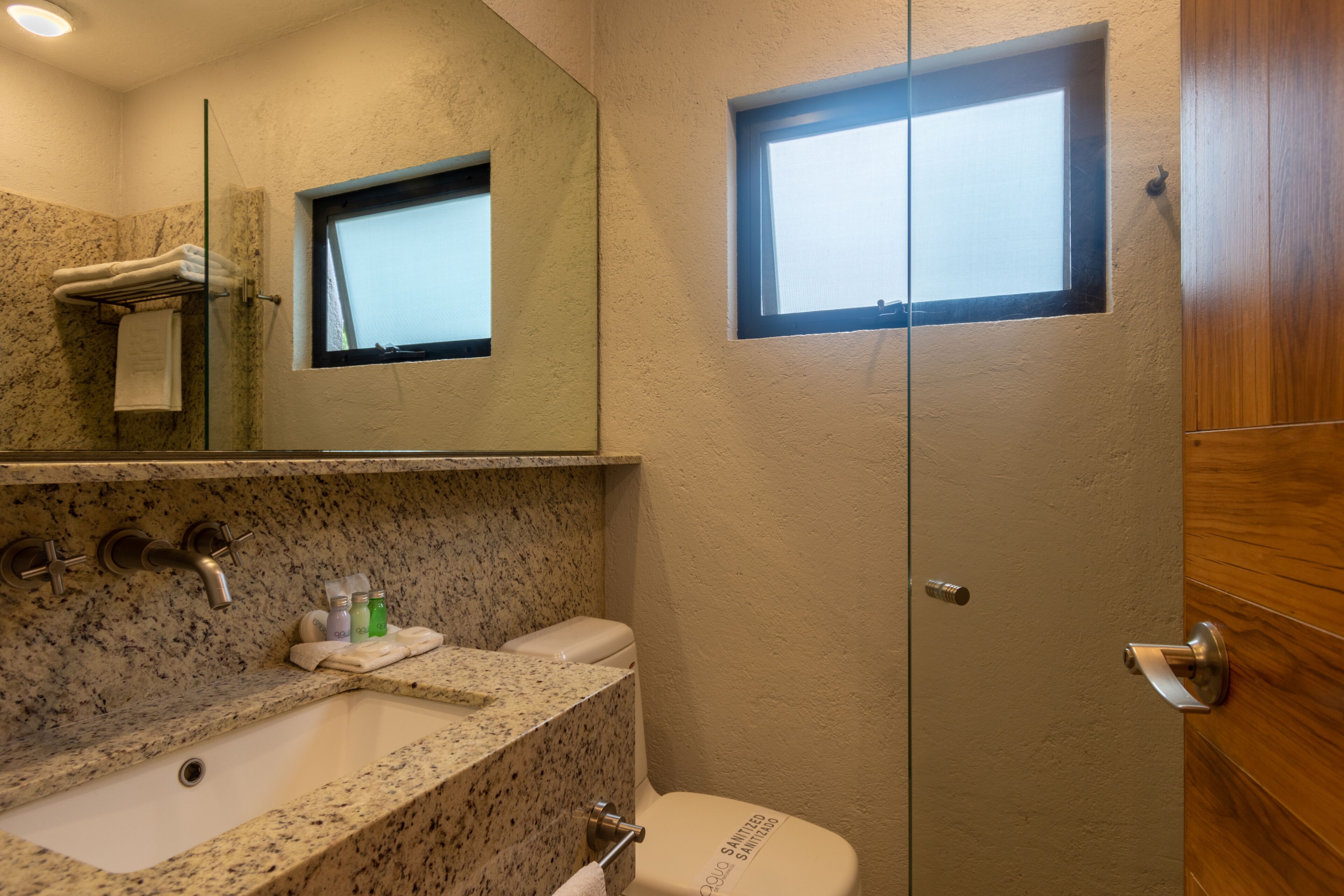 Baño con encimera de granito, lavabo, inodoro, cabina de ducha con mampara de vidrio y un ventanal.