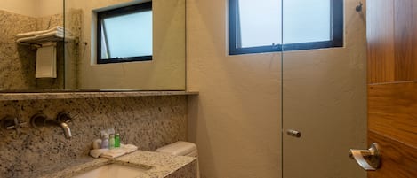 Baño con encimera de granito, lavabo, inodoro, cabina de ducha con mampara de vidrio y un ventanal.