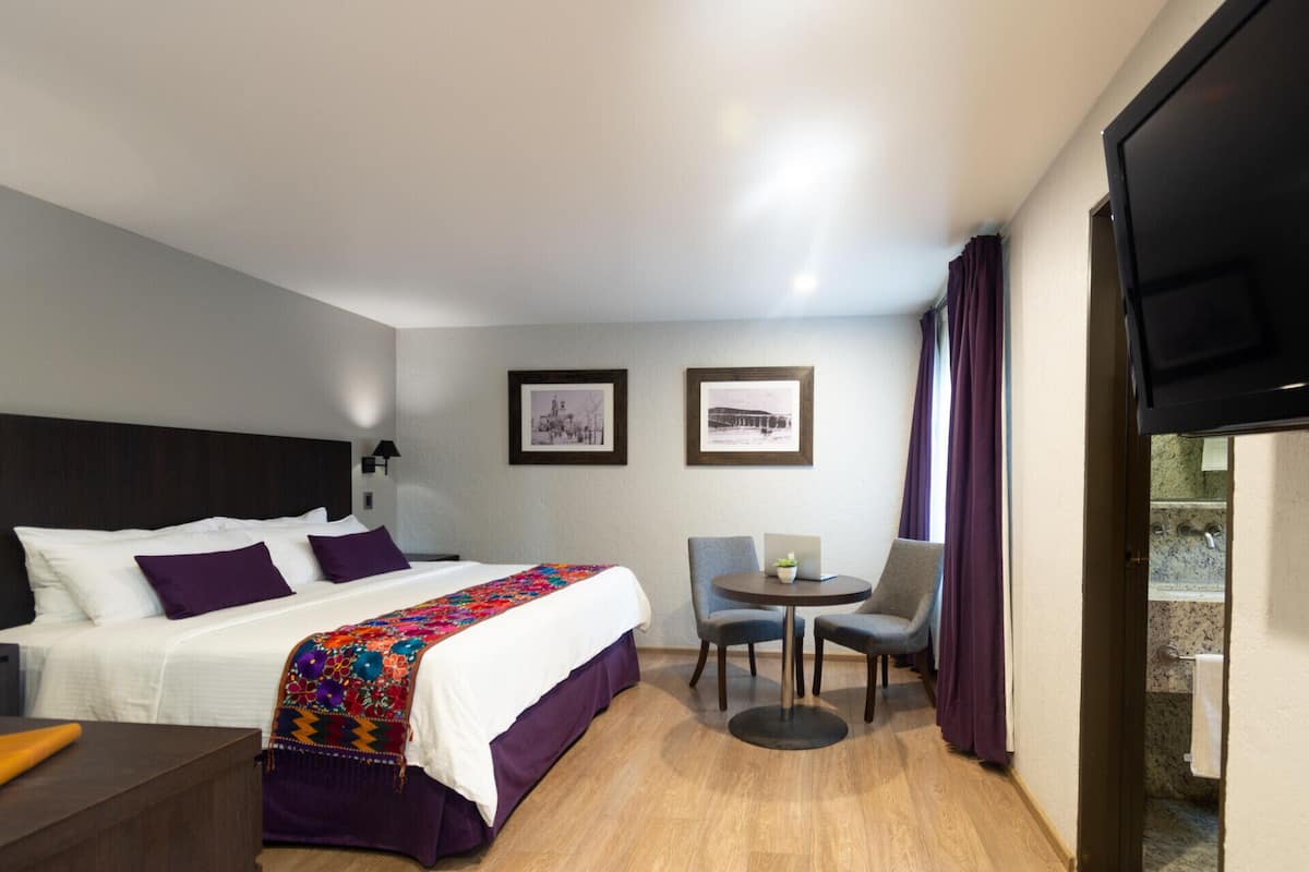 A hotel room with a bed, a desk, a chair, a small table, and two framed pictures on the wall.