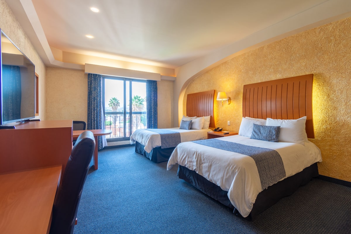 A hotel room with two beds, a desk, a chair, and a large window with curtains.