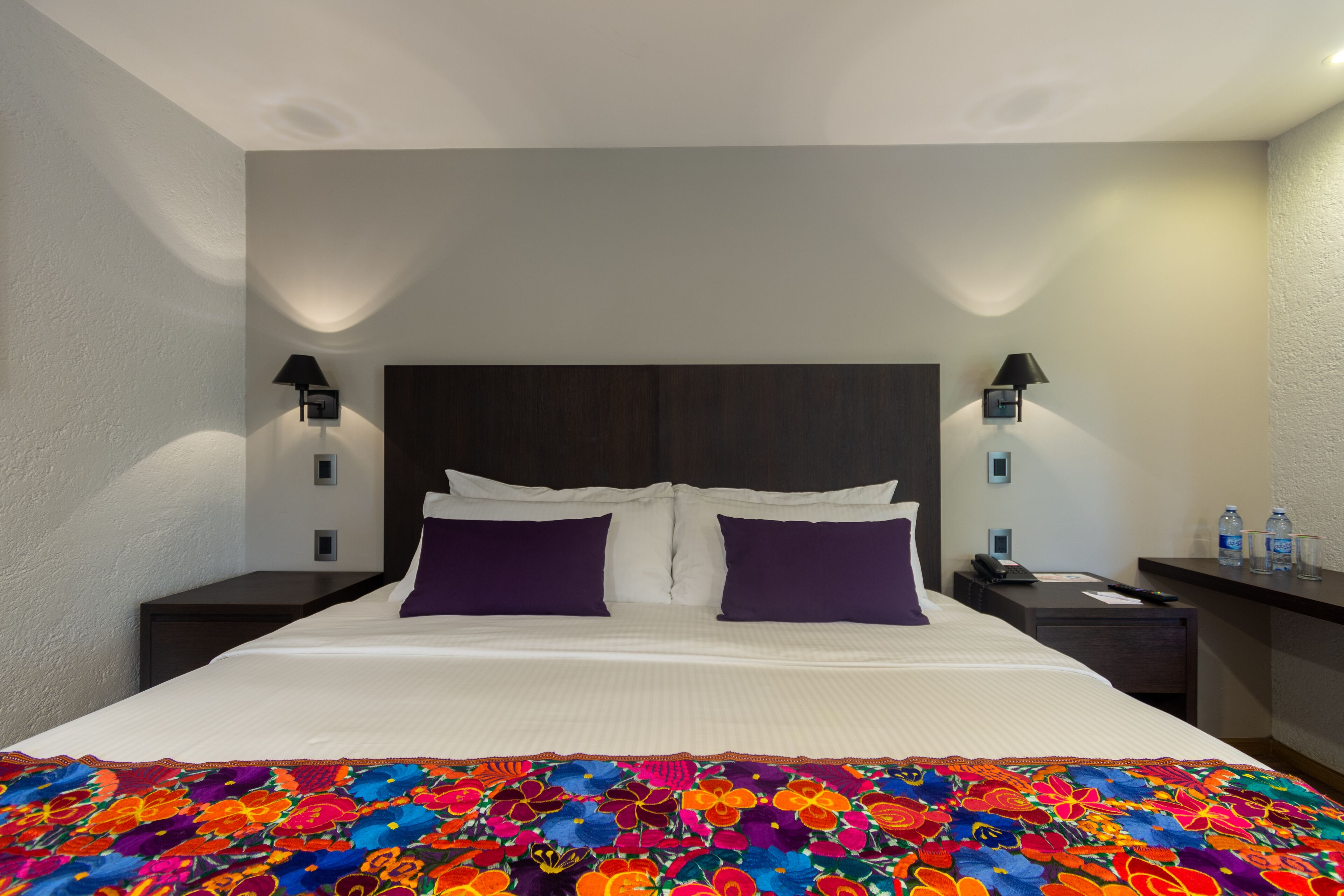 A hotel room with a large bed, two nightstands, a desk, and a night lamp on each side of the bed.