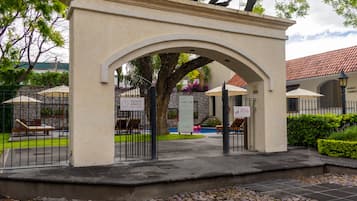 Una entrada con reja, un área de descanso techada, un muro de piedra y un letrero que indica piscina abierta las 24 horas.