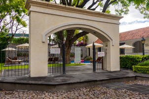 Una entrada con reja, un área de descanso techada, un muro de piedra y un letrero que indica piscina abierta las 24 horas.