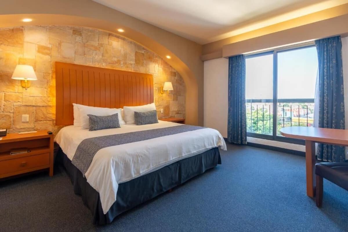 A hotel room with a stone accent wall, a wooden headboard, a large bed with a white and gray bedspread, two bedside lamps, a small table with a chair, and a large window with curtains.