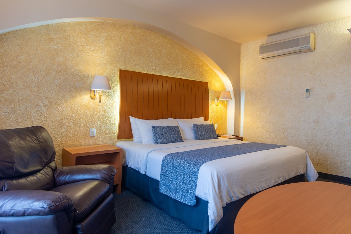 A hotel room with a bed, a leather chair, a nightstand, and a wall-mounted air conditioner.