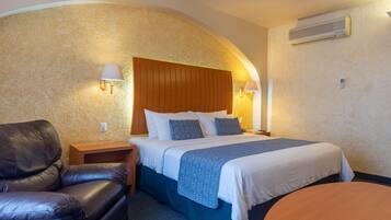A hotel room with a bed, a leather chair, a nightstand, and a wall-mounted air conditioner.