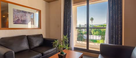 Una sala de estar moderna con un sofá de cuero, una mesa de centro de madera y una planta en maceta. Un cuadro enmarcado cuelga de la pared, y la habitación tiene un amplio ventanal corredero que da a un balcón con vistas a la vegetación y un edificio distante.