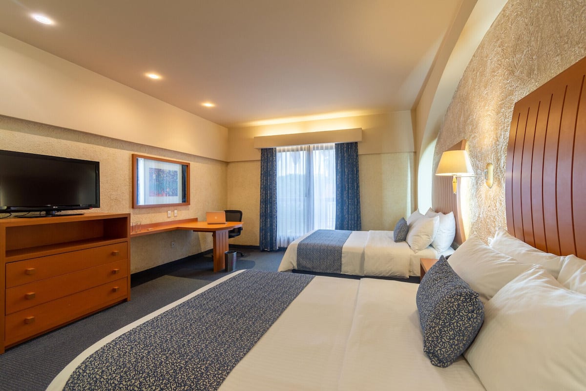 A hotel room with a TV, desk, two beds, and a window with curtains.