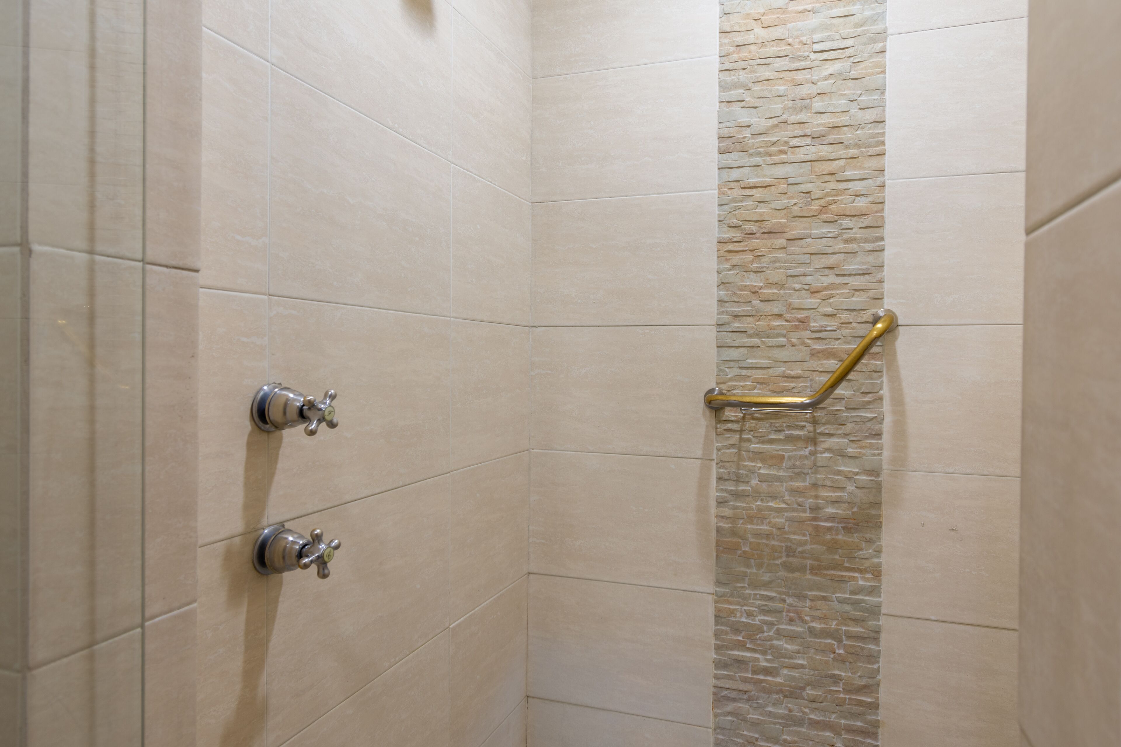 Zona de ducha con azulejos color beige, una pared de acento de piedra y un grifo de ducha dorado.