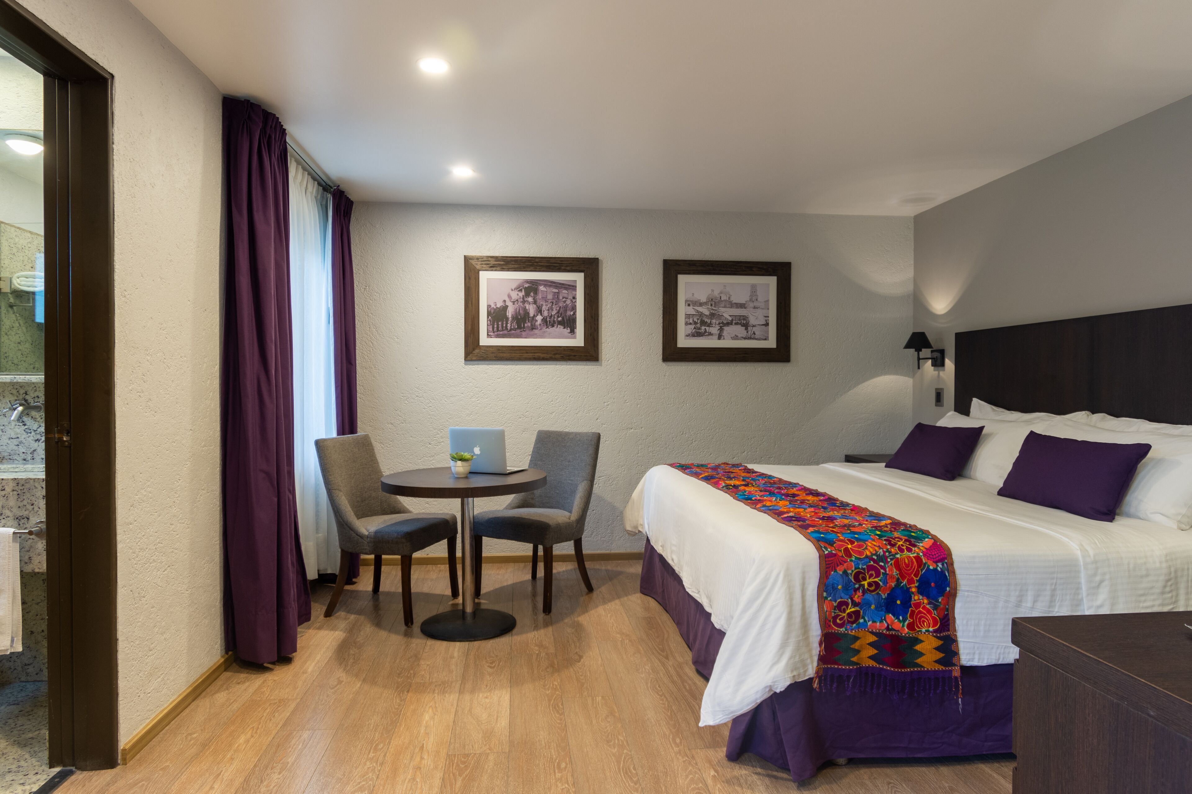 Habitación de hotel con una cama, un escritorio con una computadora portátil, dos sillas y dos cuadros en la pared.