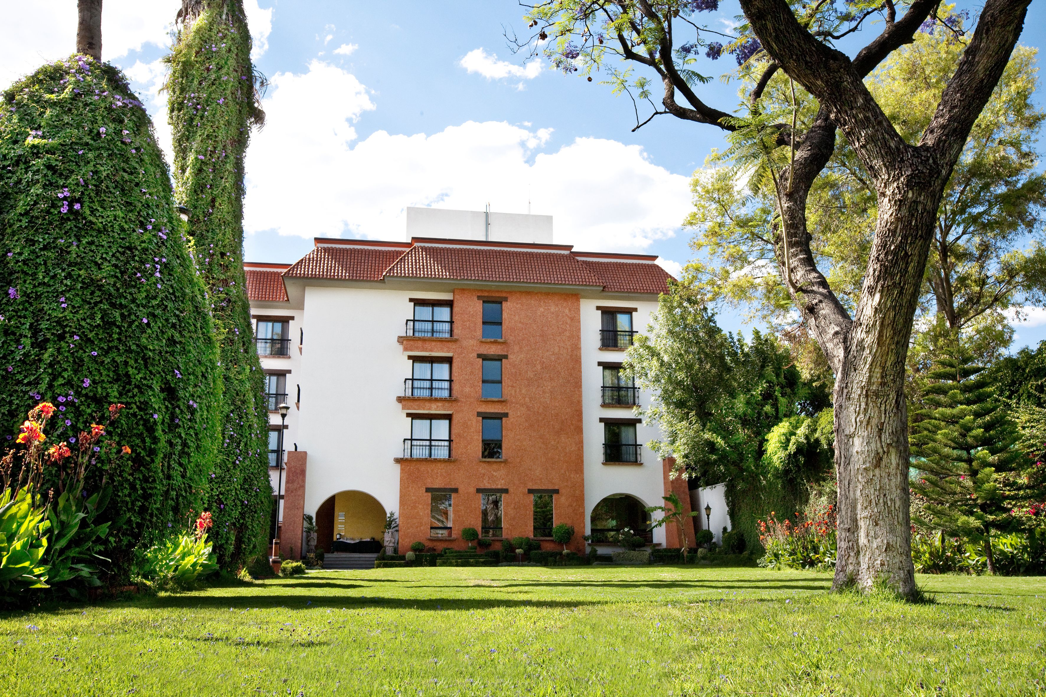 Un edificio a più piani con facciata in mattoni e livelli superiori bianchi, immerso nel verde curato e sotto un cielo sereno.