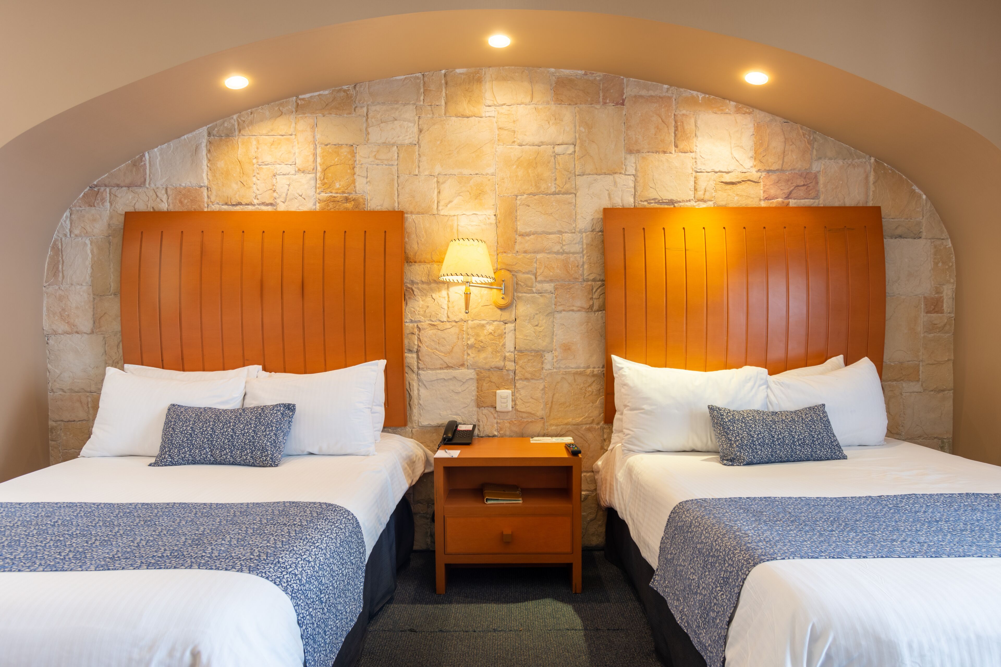 Habitación de hotel con dos camas, una pared de piedra, cabeceros de madera y una mesita de noche con una lámpara.