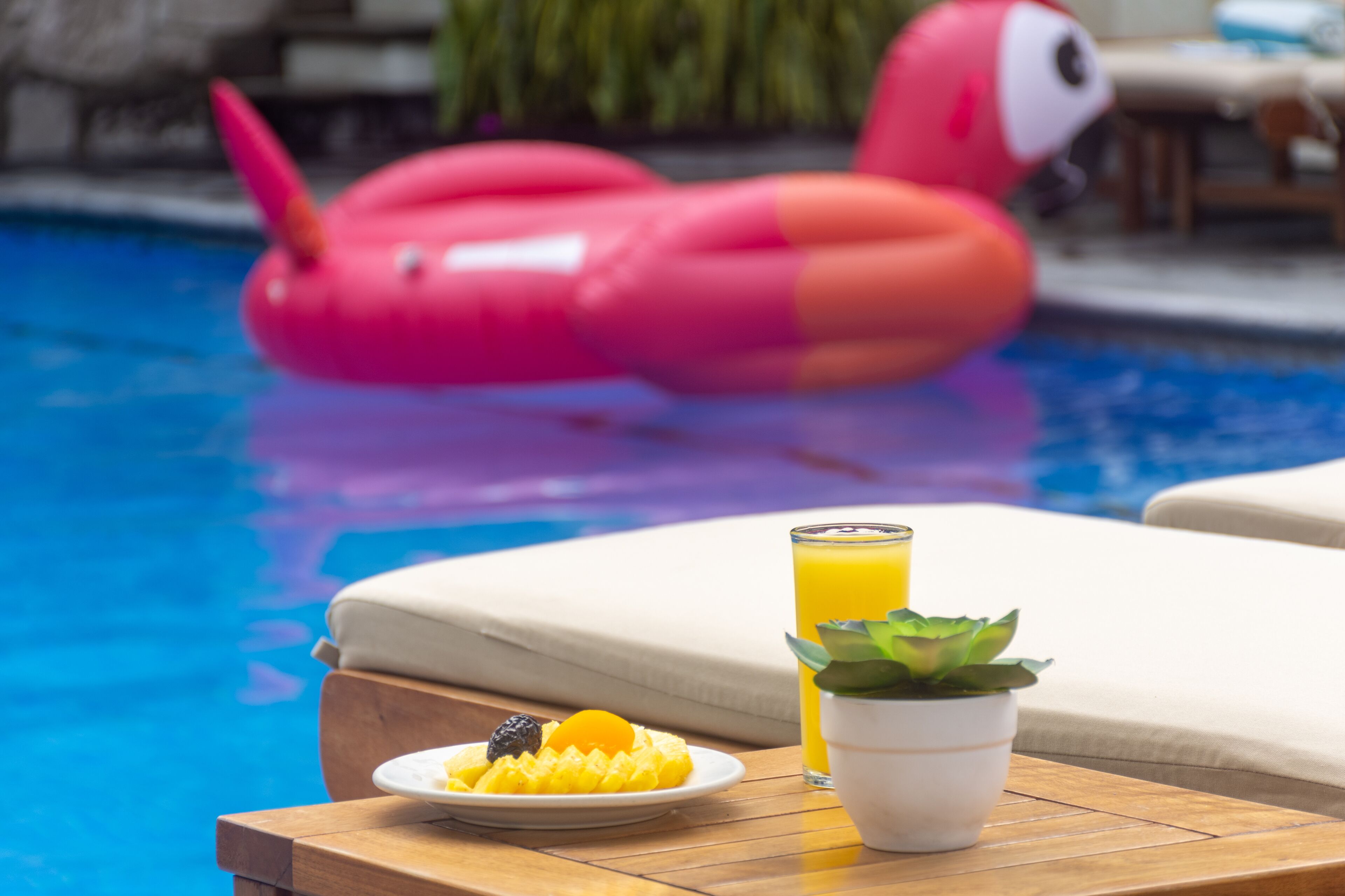 A poolside setting with a pink inflatable flamingo, a plate of fruit, a glass of orange juice, and a small potted plant on a wooden table.