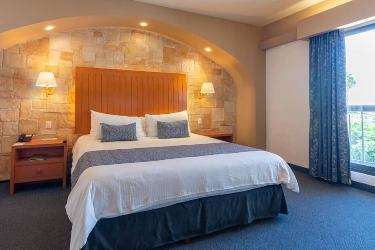 A hotel room with a stone accent wall, a wooden headboard, two bedside lamps, a bed with white bedding and two pillows, a nightstand with a phone, and a window with curtains.