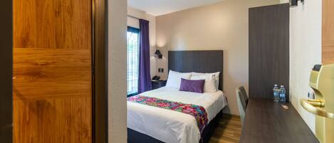 A hotel room with a wooden floor, a bed with white bedding and a colorful throw, a desk with a chair, and a window with purple curtains.